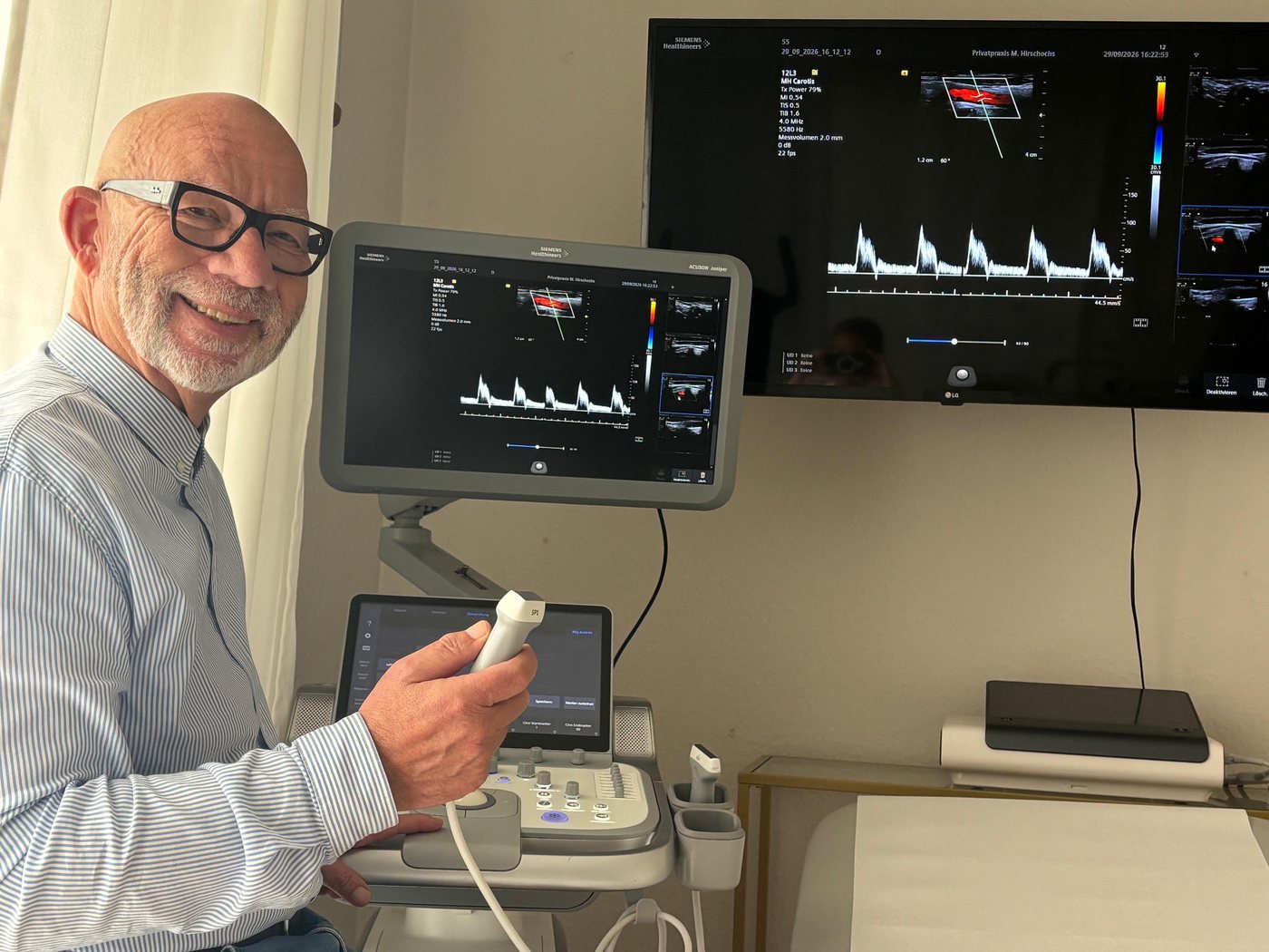 Manfred Hirschochs mit dem Schallkopf am Ultraschallgerät, auf beiden Bildschirmen eine Aufnahme der Halsschlagader im Farbdoppler